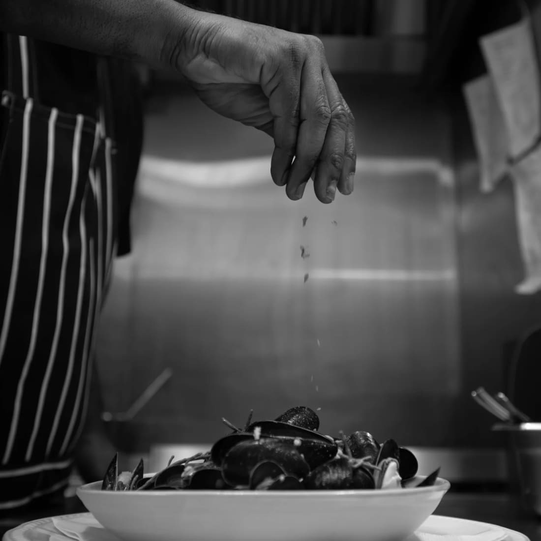 A hand seasoning a tray of mussels over the stove, in black and white
